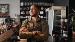 年轻快乐的非裔美国小咖啡店老板站在柜台后面，系着围裙，双臂交叉.